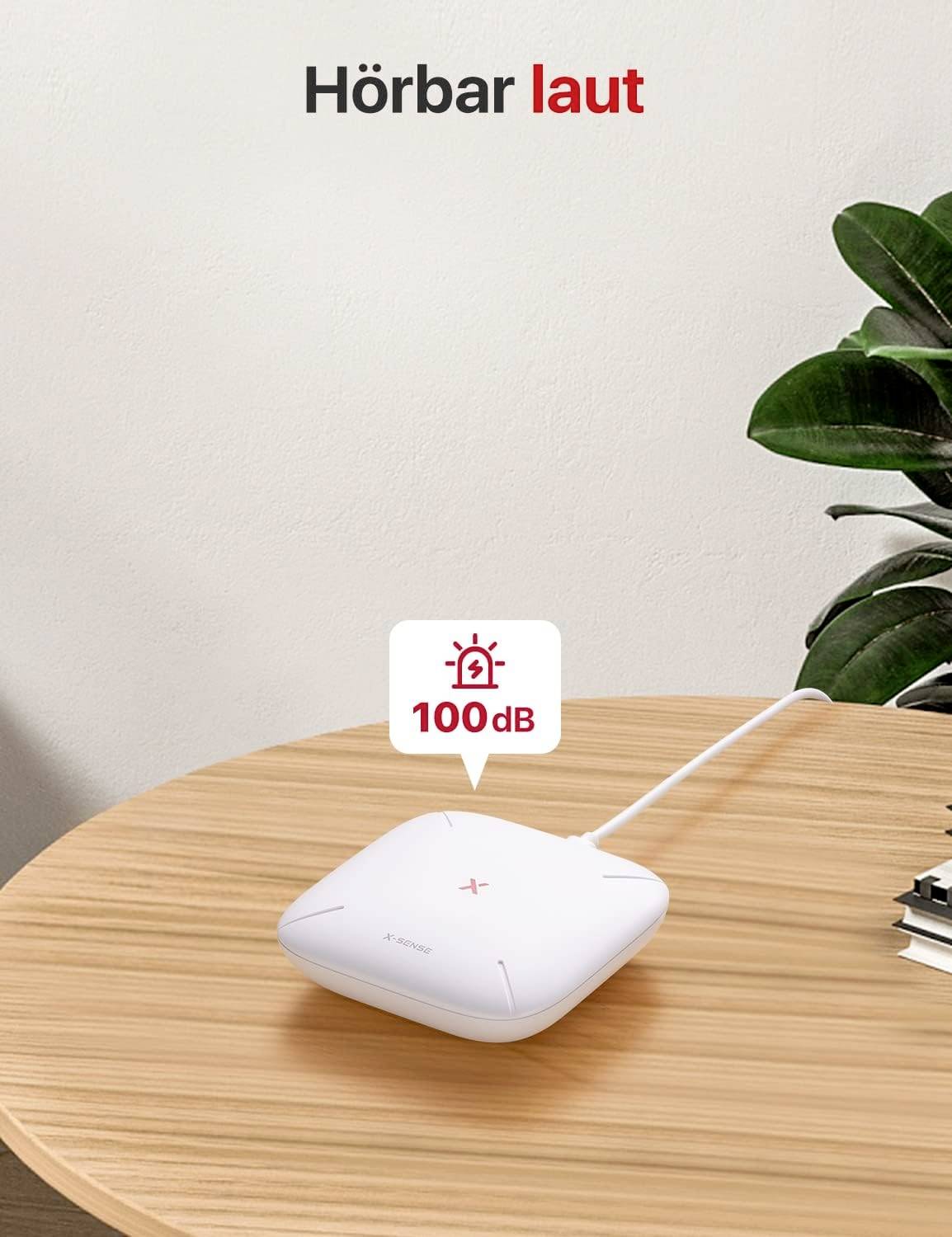 Biały alarm na drewnianym stole wskazuje 100 dB. Tekst u góry: 'Słyszalnie głośno'. Roślina doniczkowa po prawej stronie w tle.