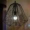 Geometryczna lampa wisząca w kształcie szkieletu z żarzącą się żarówką wisi na tle kolorowej, pikselowej ściany.