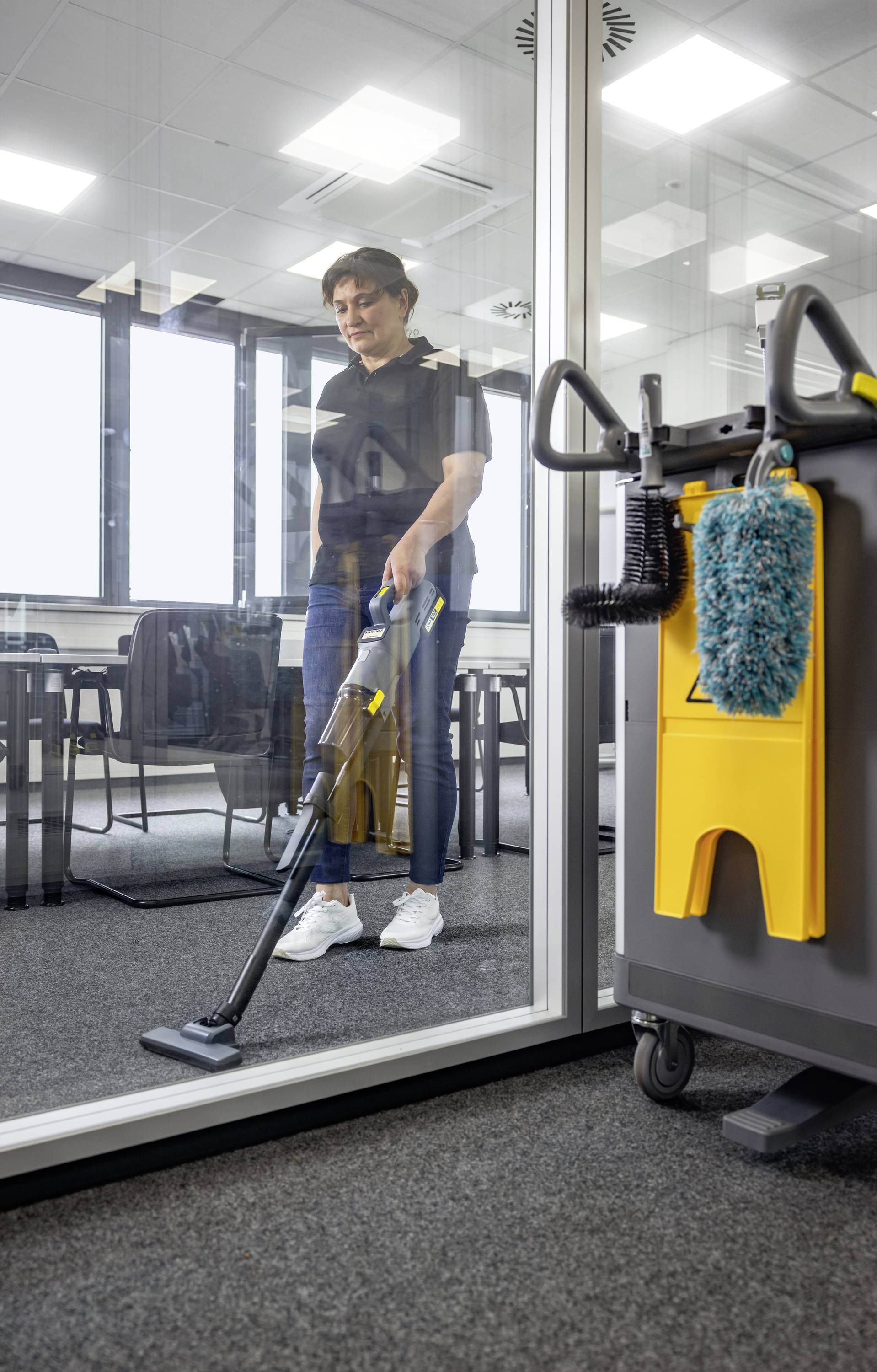 Osoba odkurzająca przestrzeń biurową wyłożoną dywanem, widoczna przez szklianą ścianę. W pobliżu znajduje się wózek sprzątający z narzędziami czystościowymi.