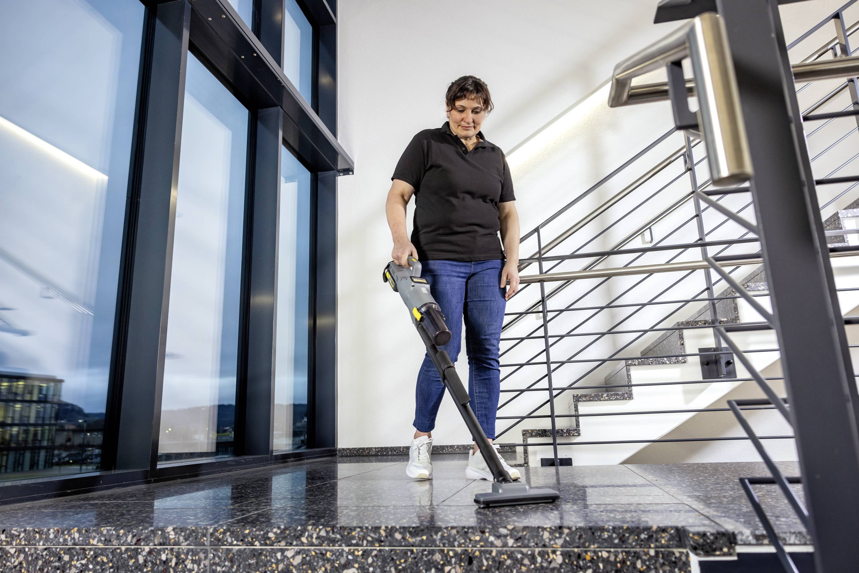 Osoba odkurza spocznik schodowy w nowoczesnym budynku za pomocą bezprzewodowego odkurzacza. Przestrzeń charakteryzuje się dużymi oknami i metalowymi poręczami.