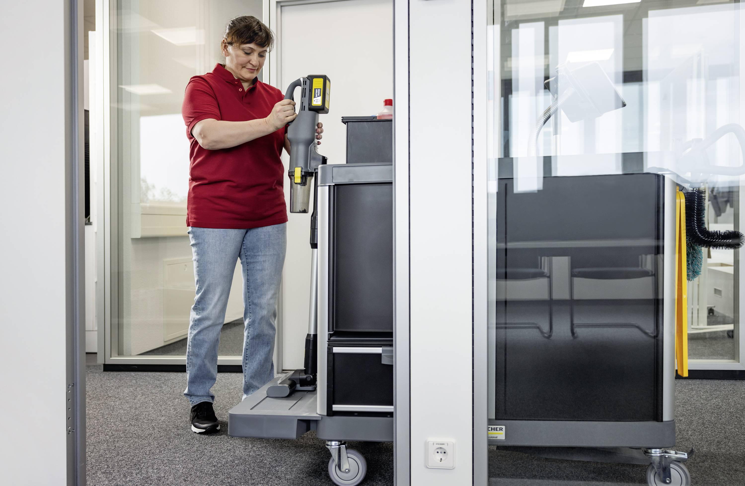 Osoba w czerwonej koszuli używa wózka sprzątającego wewnątrz biura ze szklanymi ścianami, trzymając końcówkę odkurzacza, koncentrując się na utrzymaniu czystości.