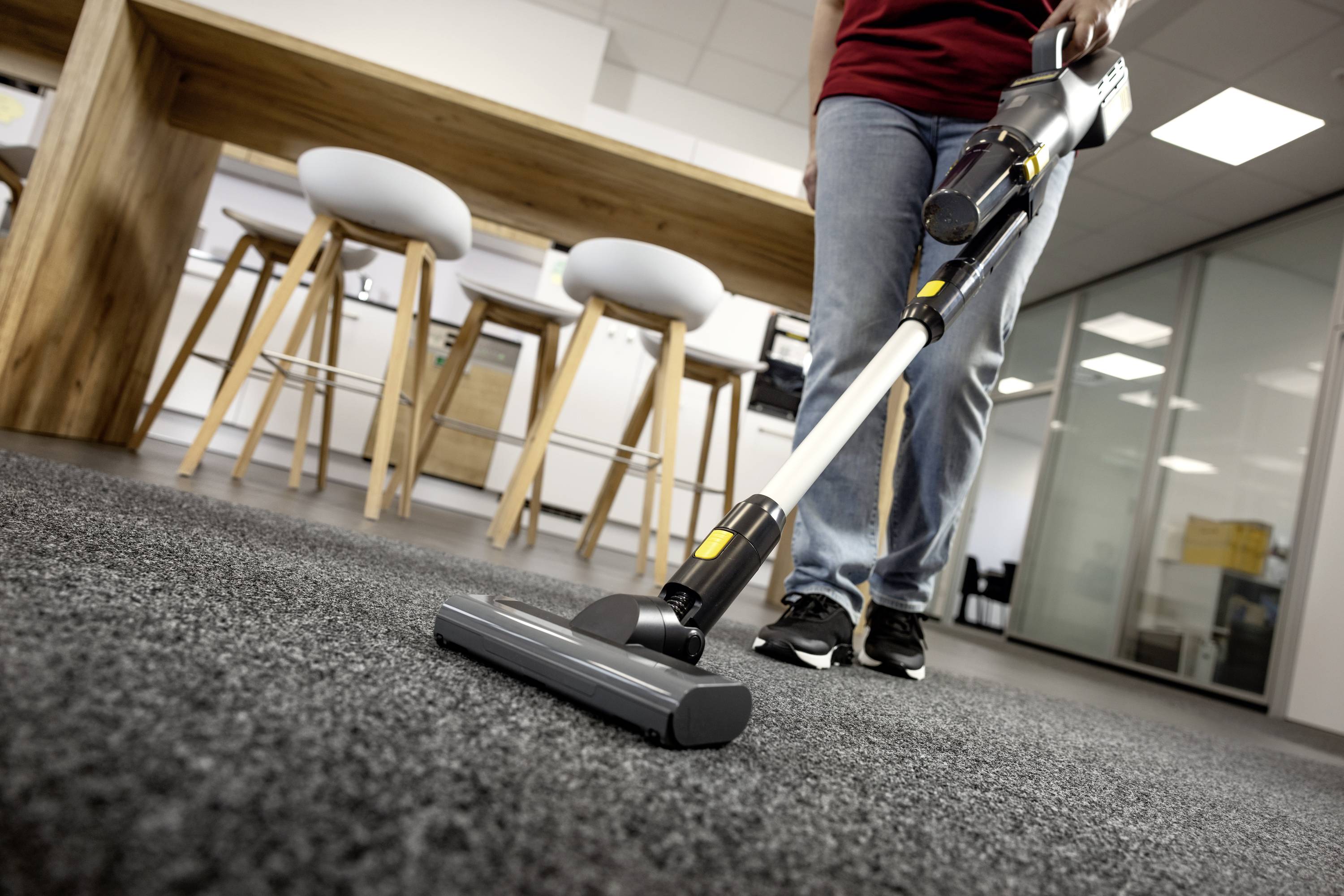 Osoba odkurzająca dywanową podłogę w nowoczesnej przestrzeni biurowej, w tle stołki i drewniany stół.