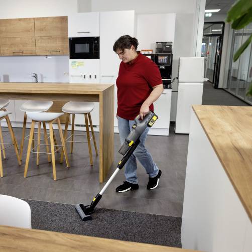 Osoba odkurza podłogę w nowoczesnej kuchni z drewnianymi szafkami i stołkami, dbając o czystość w domowym otoczeniu.