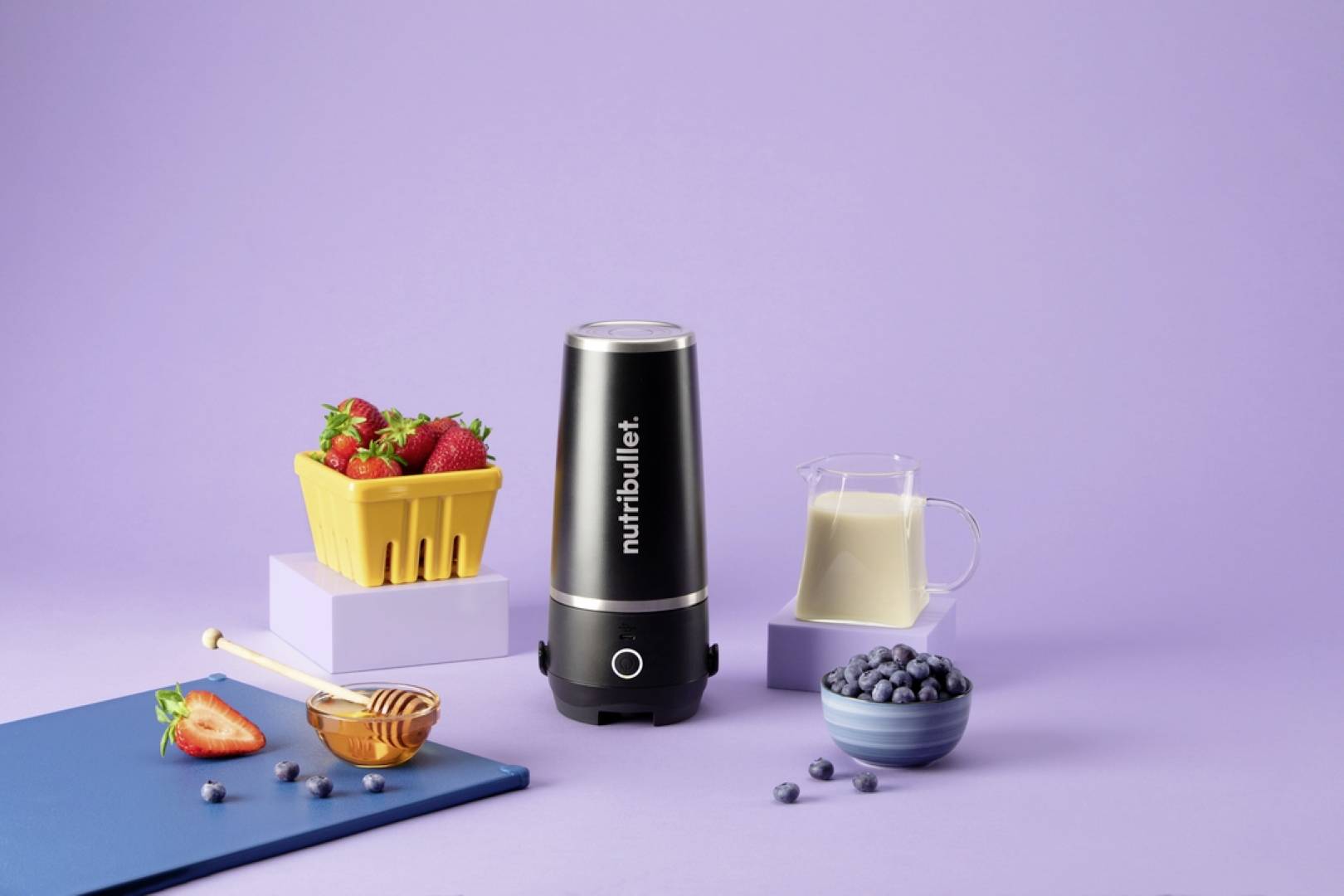 Mikser z oznakowaniem 'nutribullet' jest otoczony truskawkami, mlekiem i jagodami na fioletowym tle, sugerujących przygotowanie smoothie.