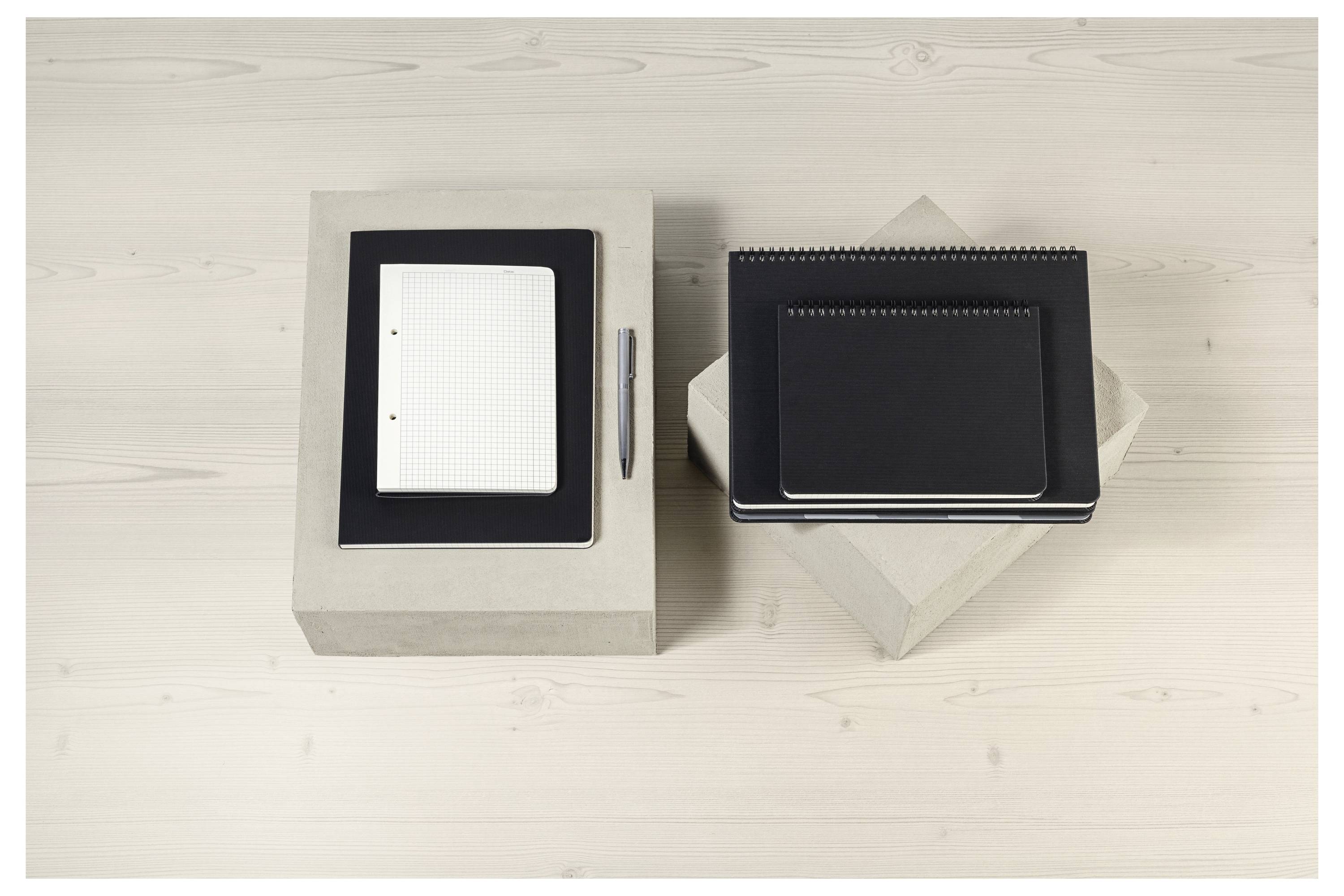 Biurko z dwoma kwadratowymi notatnikami i długopisem, ułożonymi starannie na kwadratowych podstawkach, na jasnej drewnianej powierzchni.