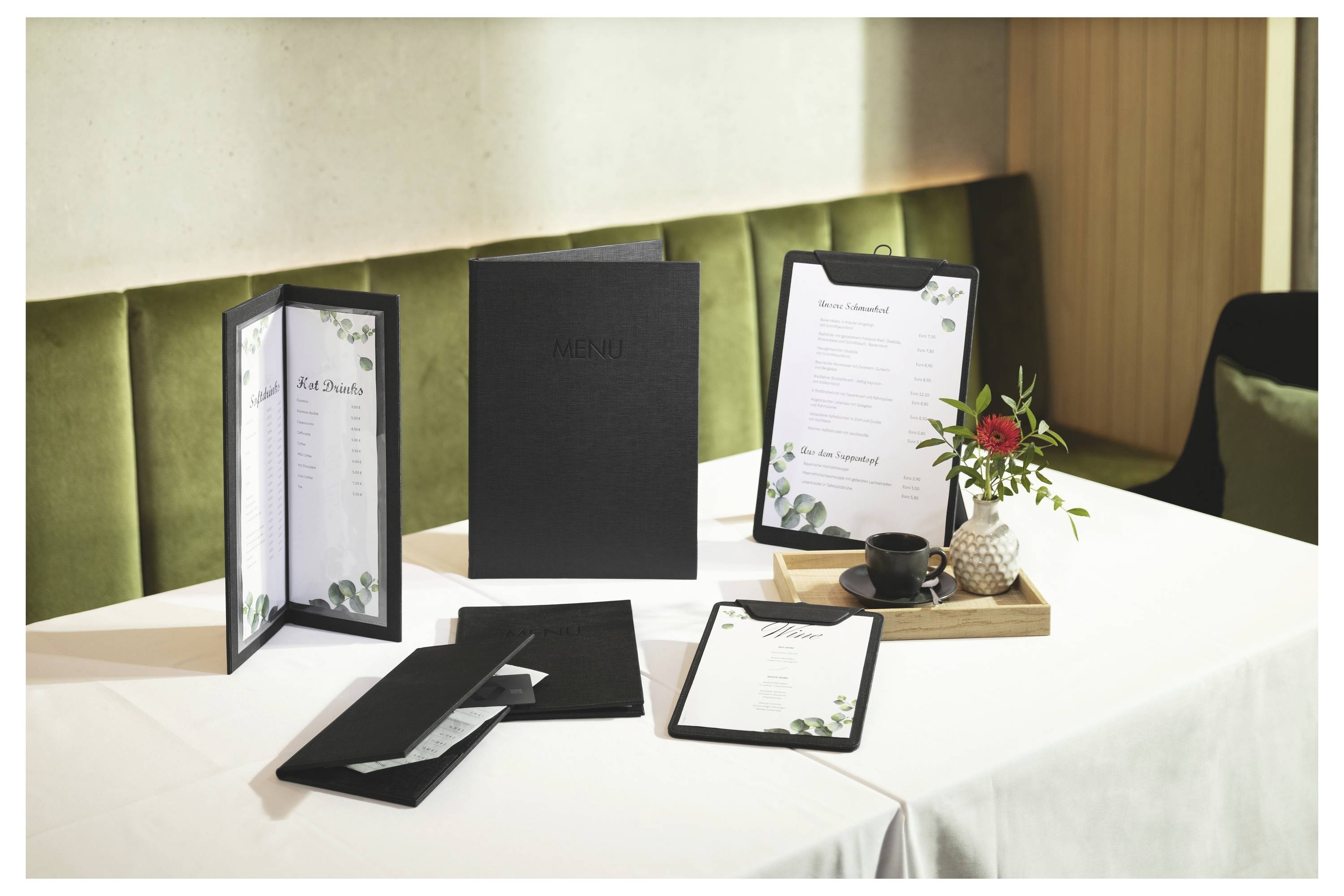 Elegancka aranżacja stołu w restauracji z czarnymi weselnym menu, złożonymi karteczkami miejsc i klipbordami florystycznymi na białym obrusie, obok małego bukietu kwiatów i filiżanki kawy.
