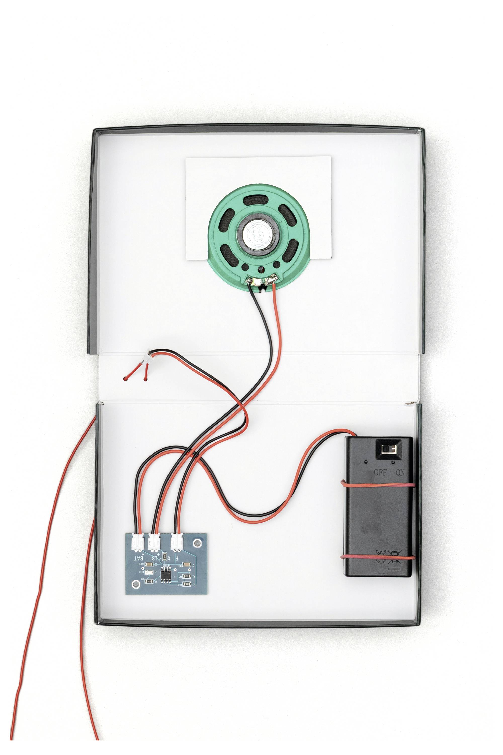 Elektroniczny zestaw z zielonym głośnikiem, płytką drukowaną, czarnym uchwytem na baterię oraz czerwonymi i czarnymi przewodami, ułożonymi w pudełku.