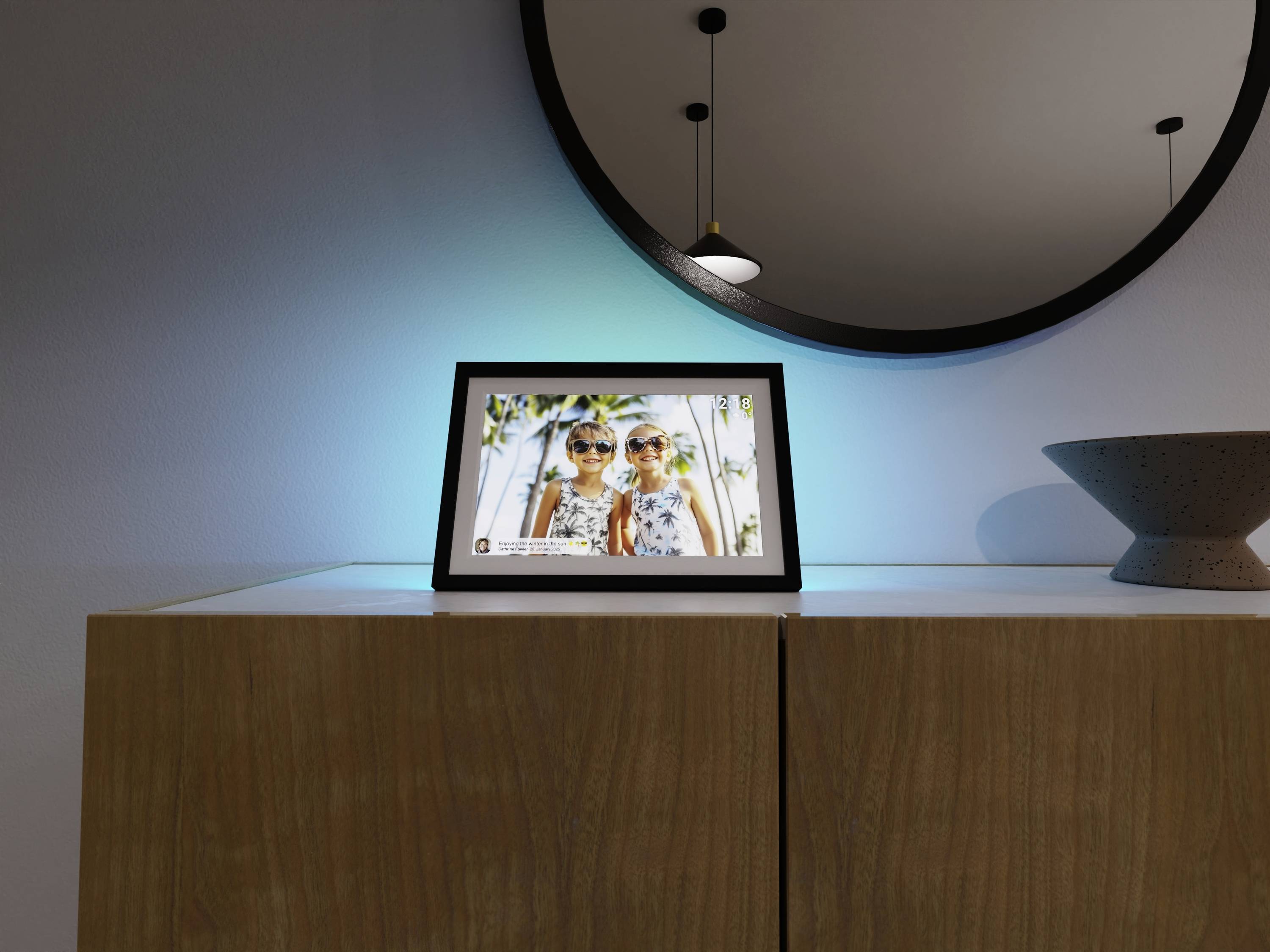 Cyfrowa ramka na zdjęcia stoi na drewnianej szafce pod okrągłym lustrem, wyświetlając zdjęcie uśmiechniętych dwojga dzieci, z nowoczesną lampą wiszącą powyżej.