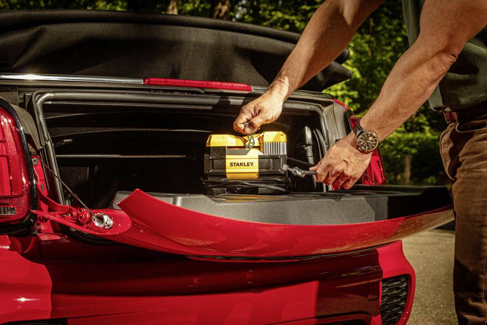 Osoba umieszcza żółte pudełko na narzędzia w bagażniku czerwonego samochodu, a w tle widać teren zalesiony.