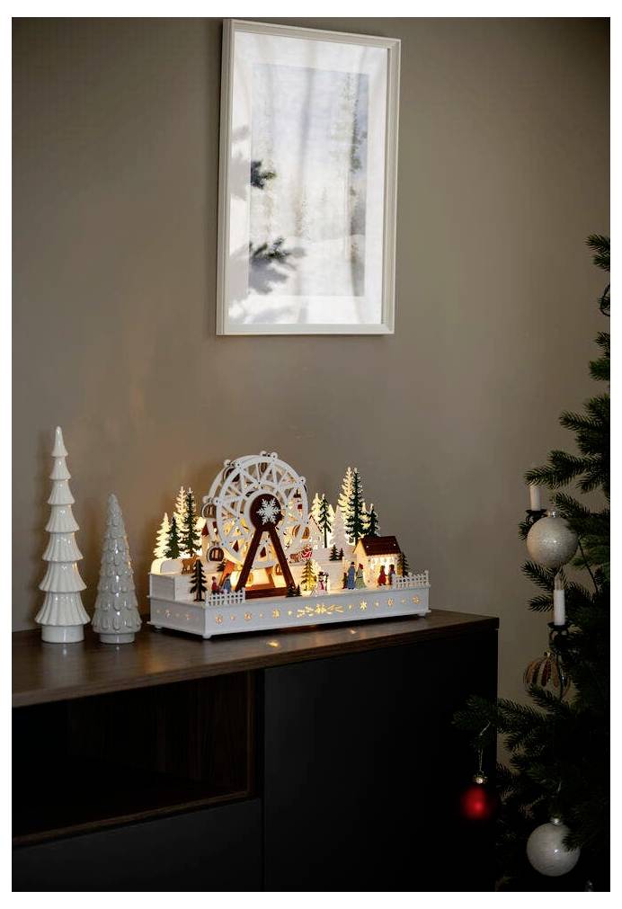 Dekoracyjna drewniana scena z podświetlonym kołem młyńskim, drzewami i domkami na szafce obok bombek choinkowych. Cienie na ściennej dekoracji.