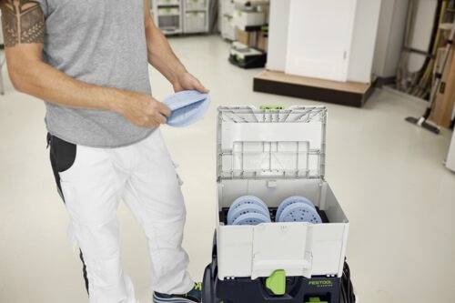 Osoba rozmieszcza okrągłe pady szlifierskie w zorganizowanym pojemniku magazynowym w warsztacie, ubrana w casualowe ubranie robocze.