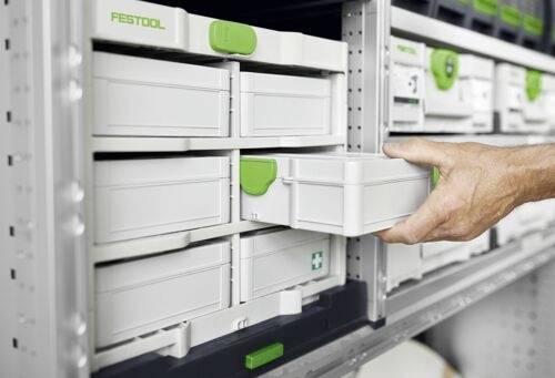 Ręka wyciąga małą szufladkę z naściennego systemu magazynowego z oznakowanymi przegródkami, podkreślając organizację i łatwość dostępu.