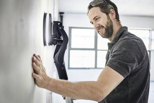 Mężczyzna używa narzędzia do wygładzania ścian w jasno oświetlonym pomieszczeniu, uśmiechając się podczas pracy.