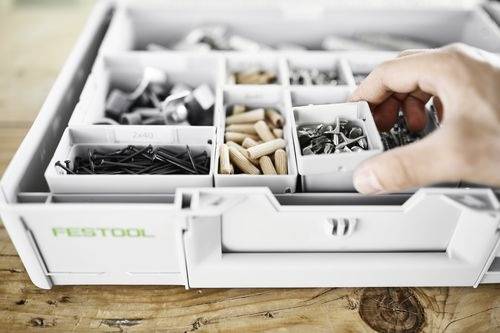 Ręka wybiera przegródkę z uporządkowanej skrzynki narzędziowej, zawierającej różne śruby, gwoździe i drewniane kołki, stojącej na drewnianym stole.