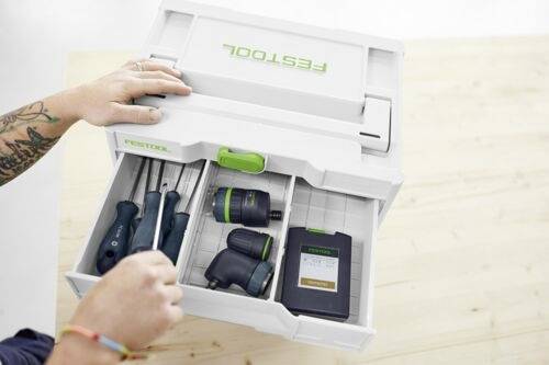 Osoba rozmieszcza narzędzia w skrzynce magazynowej Festool, w tym wkrętaki, wiertarkę i baterię, na jasnej drewnianej powierzchni.