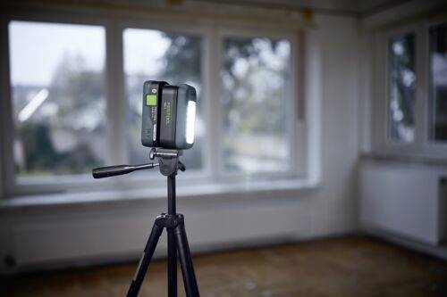 Statyw z zamontowanym aparatem fotograficznym i jasnym światłem stoi w pustym pokoju z dużymi oknami, co sugeruje koncentrację na fotografii lub nagrywaniu.