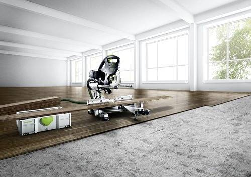 Piła elektryczna leży na niewykoańczonej podłodze w jasnym pomieszczeniu. Drewniane deski i skrzynka narzędziowa znajdują się w pobliżu, sugerując trwające prace remontowe.