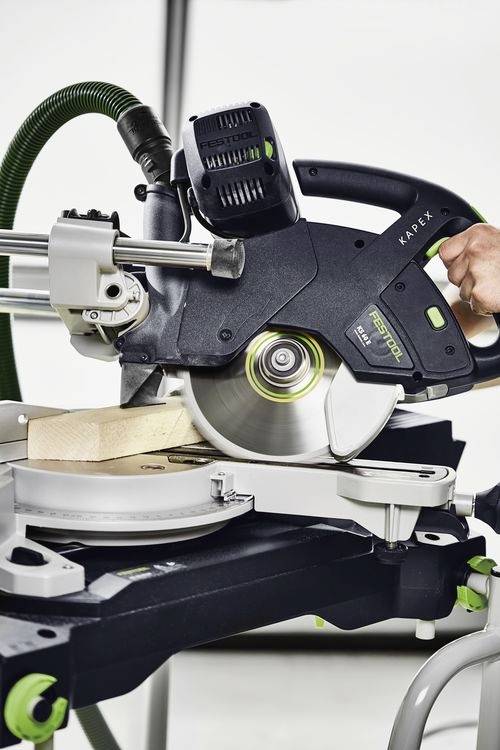 Osoba obsługuje piłę ukośnicę, precyzyjnie przecinając kawałek drewna. Piła jest czarno-srebrna z zielonymi akcentami, zamocowana do stołu warsztatowego.
