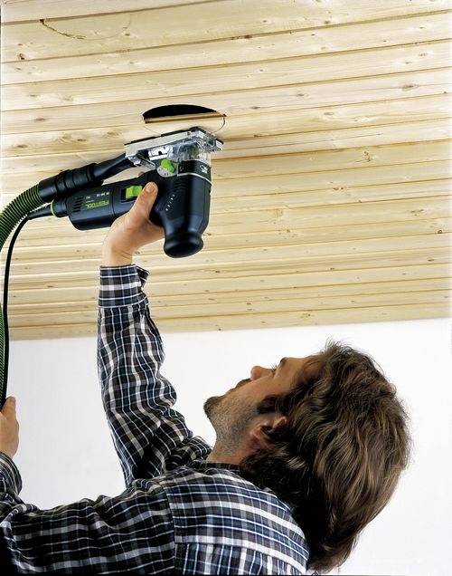 Osoba używa elektronarzędzia do wywiercenia otworu w drewnianym suficie, a narzędzie jest podłączone do węża odprowadzającego pył.