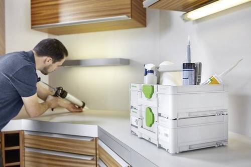 Osoba nakłada uszczelniacz wzdłuż narożnika blatu kuchennego za pomocą pistoletu do uszczelniania; środki czyszczące są widoczne na blacie.