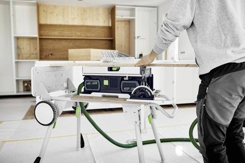 Osoba pracująca przy pile tarczowej, która ucina drewnianą deskę w jasno oświetlonym pomieszczeniu z półkami. Na stole widoczne są narzędzia bezpieczeństwa.