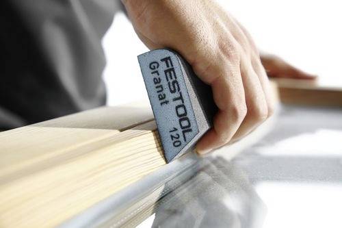 Dłoń szlifująca drewnianą powierzchnię za pomocą bloku szlifierskiego Festool Granat 120, podkreślająca rzemiosło stolarskie i wykańczanie powierzchni.