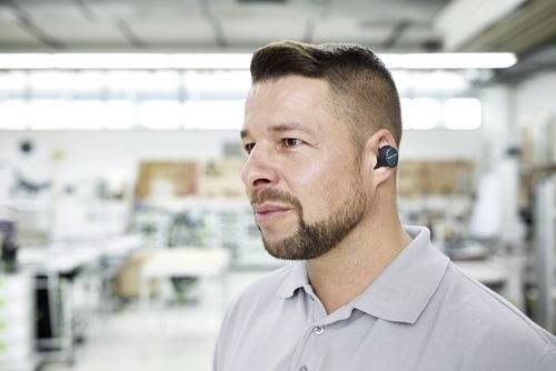 Osoba ubrana w szarą koszulę z kołnierzykiem i bezprzewodowy słuchawka douszną stoi w jasnej, nowoczesnej przestrzeni pracy. Tło jest rozmyte i przemysłowe.