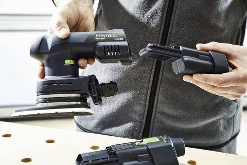 Osoba trzyma bezprzewodową szlifierkę Festool oraz dwie baterie na drewnianym warsztacie, co wskazuje na wymienne źródła zasilania.