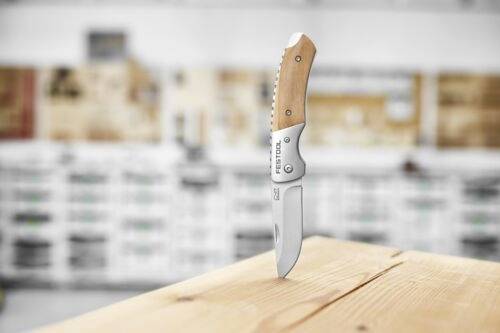 Składany nóż z drewnianą rękojeścią i ostrzem ze stali nierdzewnej stoi pionowo na drewnianej powierzchni, na rozmytym tle warsztatu.