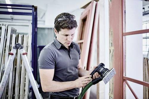 Osoba w warsztacie używa elektronarzędzia do szlifowania drewnianej ramy, otoczona różnymi elementami okiennymi i sprzętem.