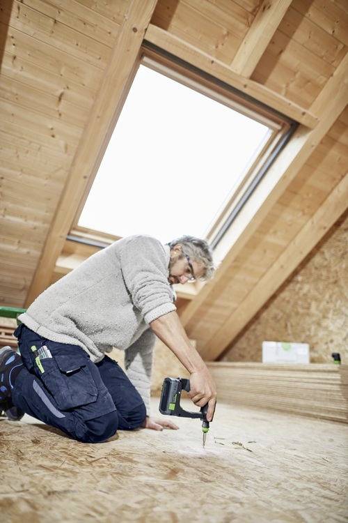 Osoba klęczy na drewnianej podłodze pod świetlikiem, używając wiertarki elektrycznej. Pomieszczenie wydaje się być poddaszem lub przestrzenią poddasza w trakcie remontu.