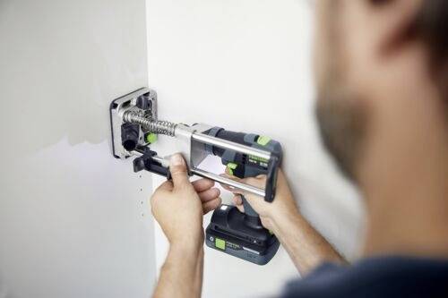 Osoba używa wiertarki elektrycznej do zamontowania metalowego zawiasu drzwiowego na białej powierzchni, koncentrując się na precyzji i wyrównaniu.