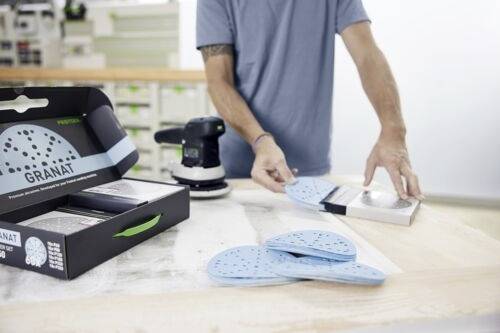 Osoba w warsztacie układa niebieskie tarcze szlifierskie oznaczone napisem "GRANAT" obok szlifierki na stole warsztatowym.