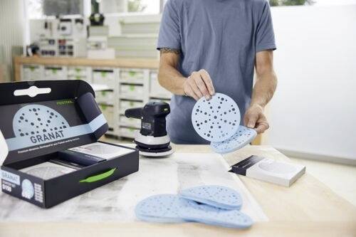 Osoba prezentuje kilka niebieskich krążków ściernych w pobliżu szlifierki stacjonarnej na warsztacie. Na stole znajduje się pudełko oznaczone napisem "Granat".