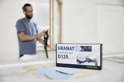 Pudełko oznaczone etykietą "Granat D125 Zestaw Startowy" z tarczami szlifierskimi leży na stole. W tle, nieostro, widać osobę trzymającą narzędzie.