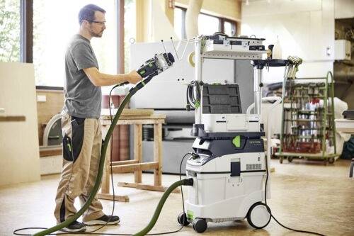 Osoba używająca przemysłowego systemu odkurzającego w warsztacie, obsługująca wąż podłączony do jeżdżącej jednostki z panelem sterowania.