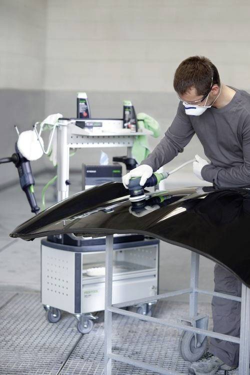 Osoba w masce i rękawiczkach używa maszyny do polerowania czarnej pokrywy silnika samochodu w warunkach warsztatu.