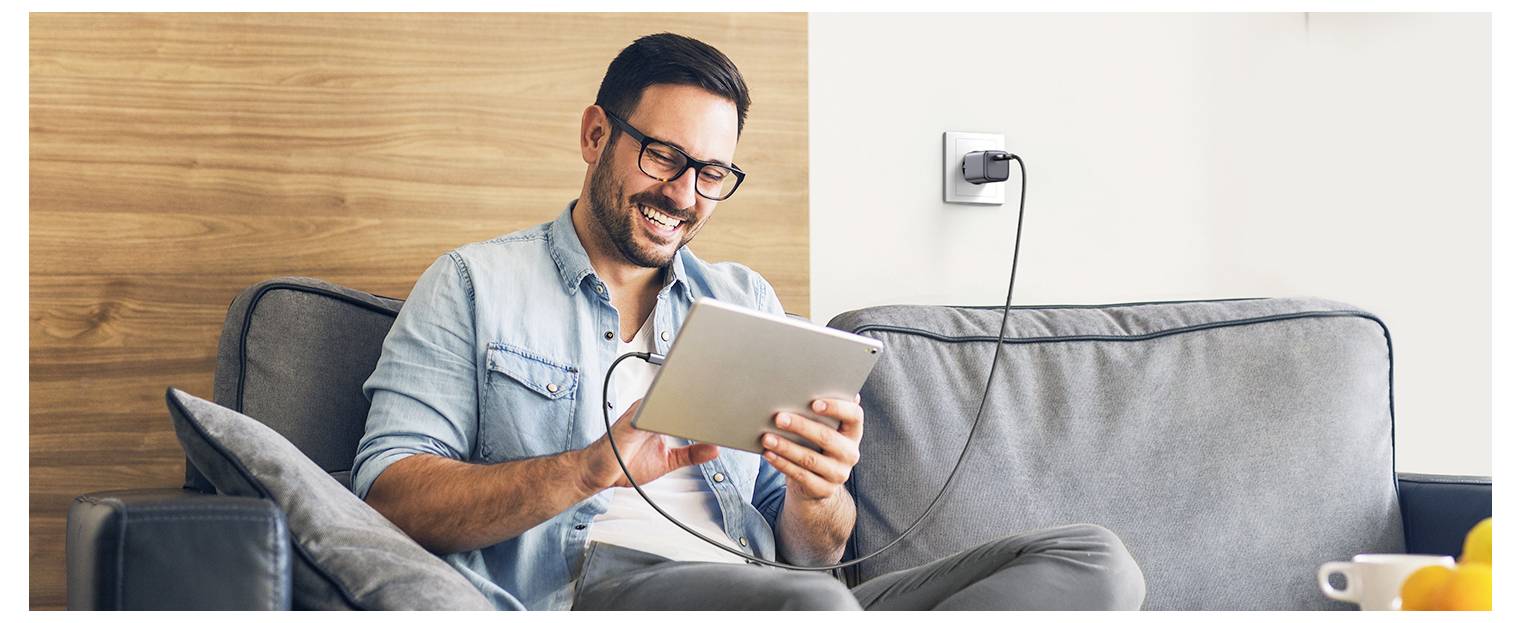 Mężczyzna siedzący na kanapie w luźnej pozie, uśmiecha się podczas korzystania z tabletu podłączonego do gniazdka elektrycznego do ładowania.