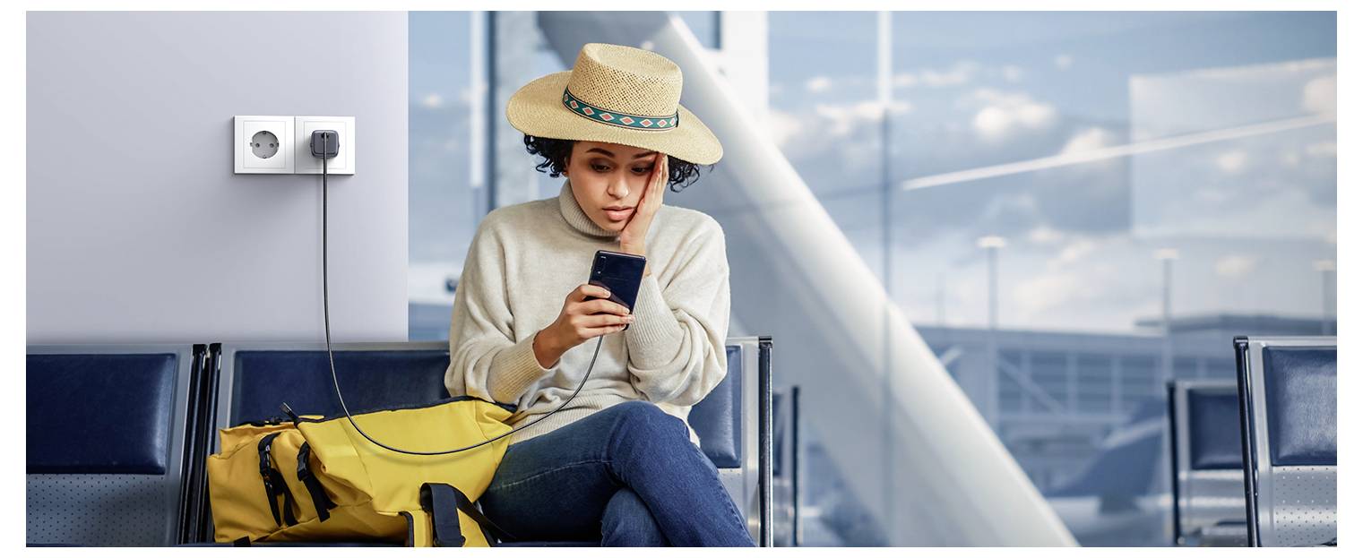 Osoba w słomkowym kapeluszu ładuje telefon w poczekalni lotniska, siedząc na ławce obok żółtego plecaka i gniazdka elektrycznego.