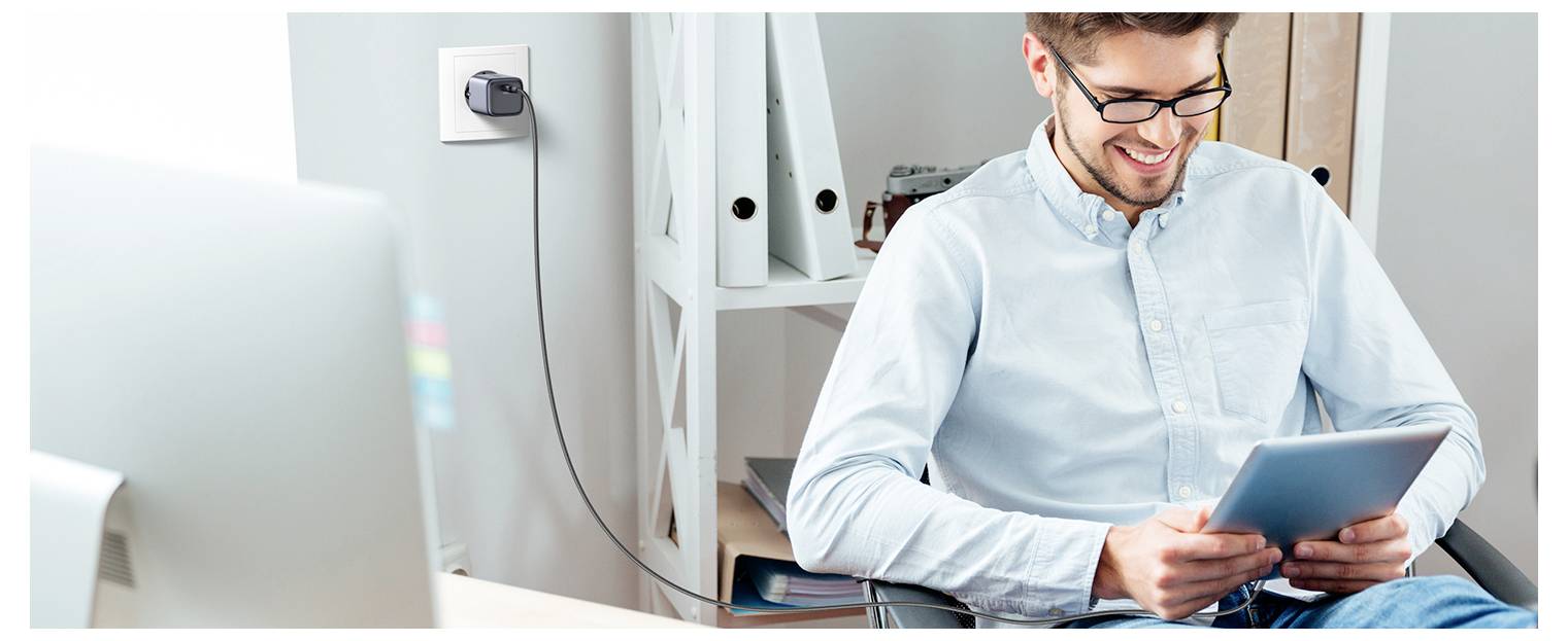 Osoba siedzi w biurze, uśmiechając się do tabletu. Na pierwszym planie widać biurko z komputerem i półki z segregatorami w tle, co sugeruje środowisko pracy lub nauki.