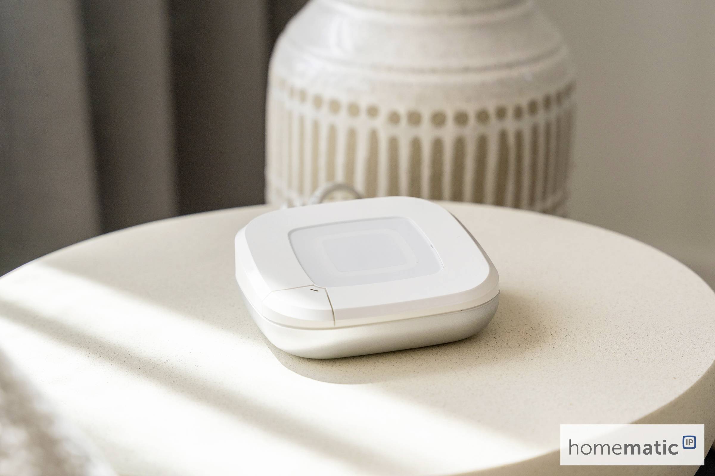 Mały biały urządzenie do automatyzacji domu spoczywa na okrągłym beżowym stole w miękko oświetlonym pomieszczeniu. W rogu widoczne jest logo 'homematic'.