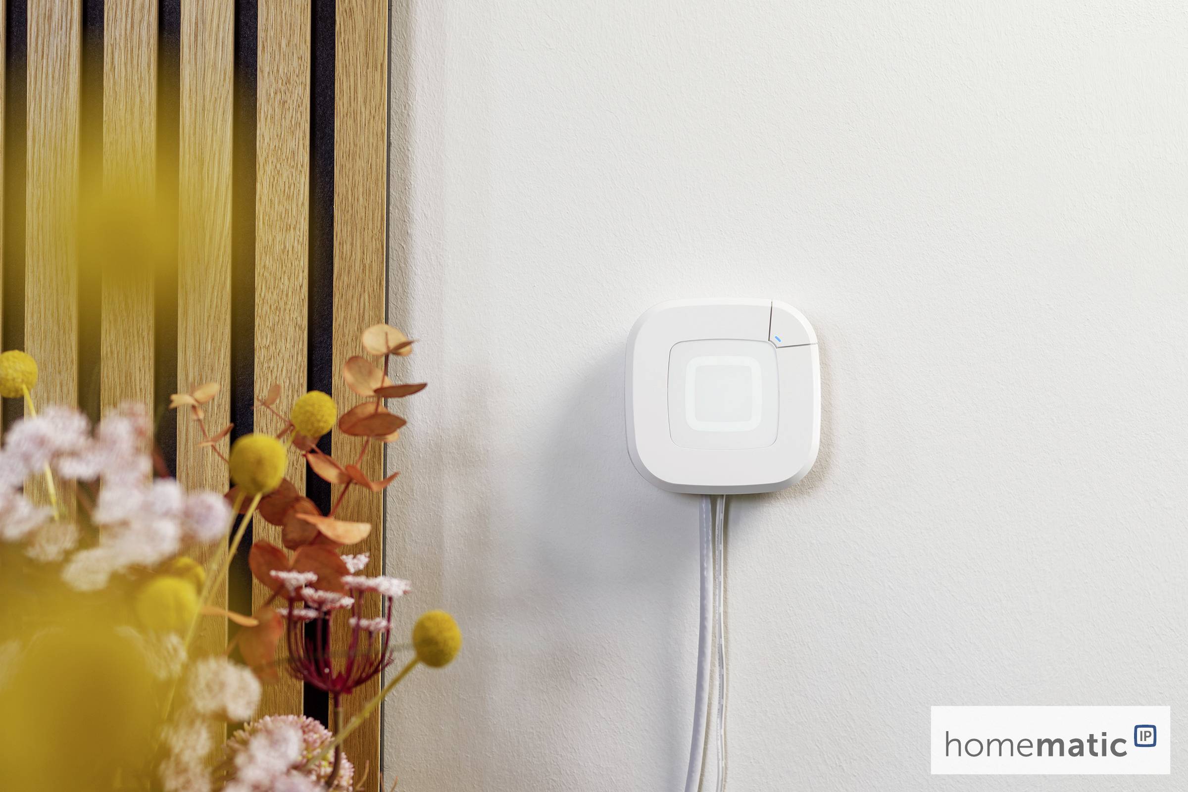 Bezprzewodowe urządzenie inteligentnego domu zamontowane na ścianie obok dekoracyjnych kwiatów, z podświetlonym wskaźnikiem.