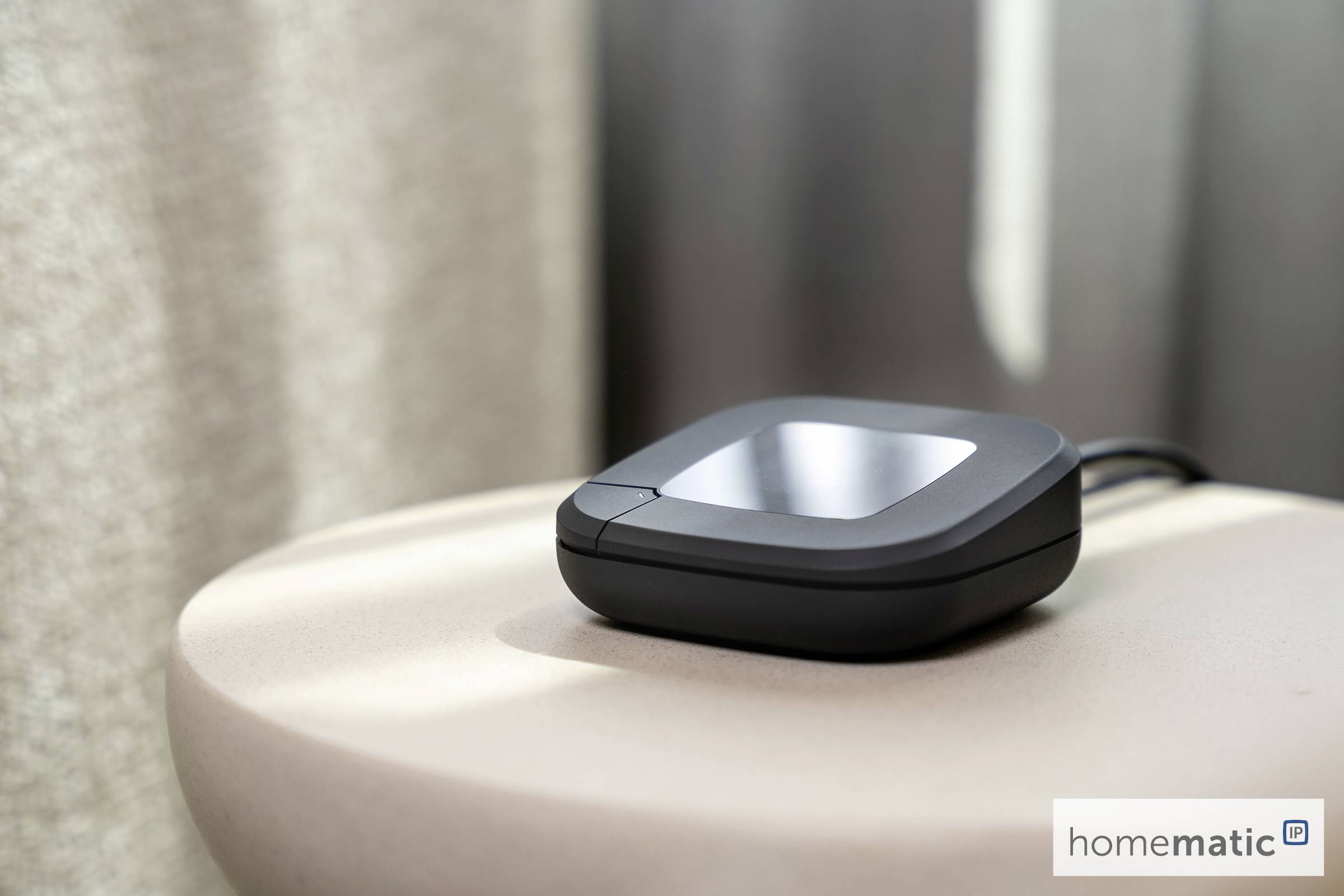 Mały, czarny, kwadratowy elektroniczny przyrząd leży na okrągłym, jasnym stole, z podłączonym przewodem. Etykieta głosi 'homematic.'