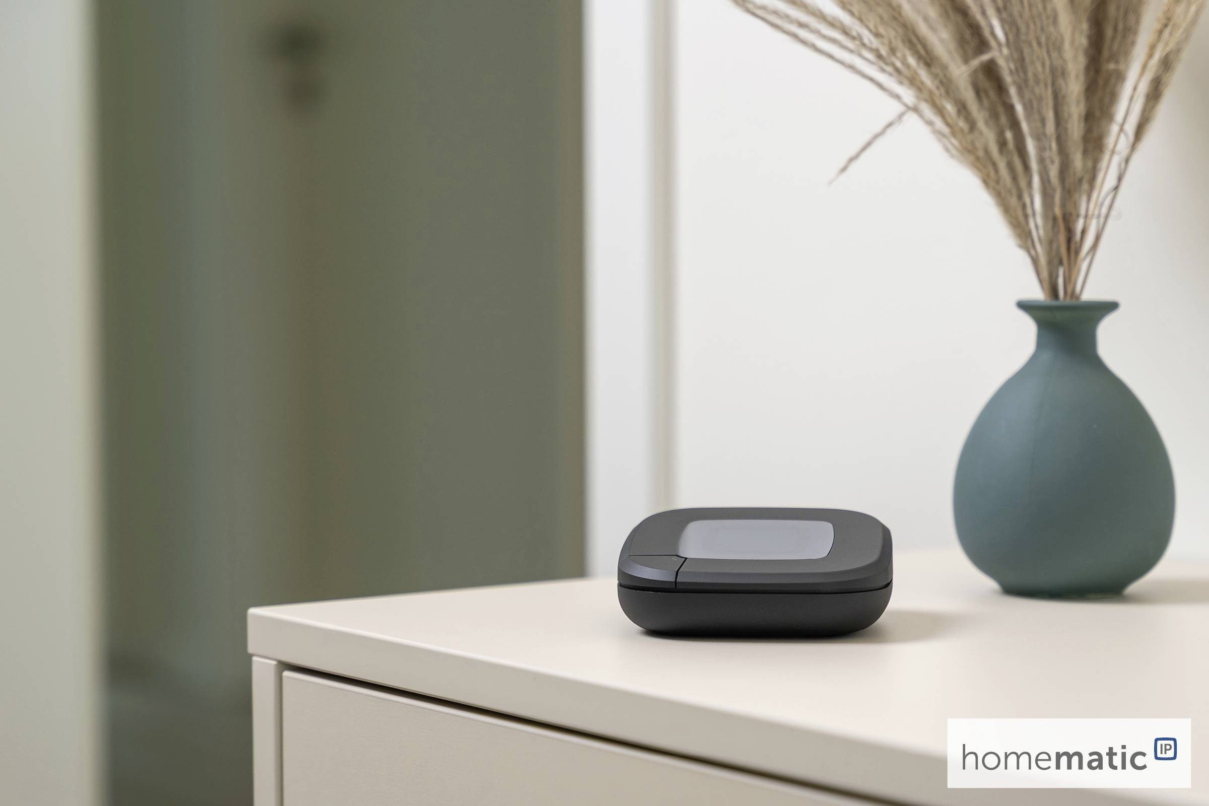 Mały czarny urządzenie z wyświetlaczem cyfrowym stoi na jasnobiałym stole obok turkusowego wazonu z suszoną trawą. Logo na urządzeniu głosi 'homematic IP'.
