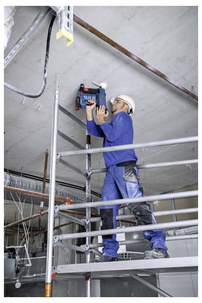Robotnik w niebieskim kombinezonie i kasku ochronnym stoi na rusztowaniu, używając elektronarzędzia do wiercenia w suficie na placu budowy.