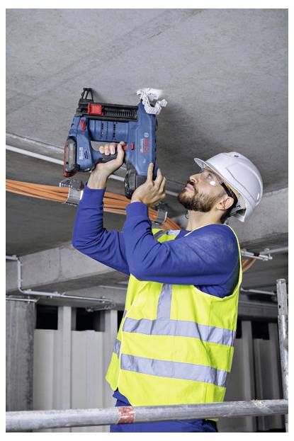 Robotnik budowlany w kasku ochronnym i kamizelce odblaskowej używa elektronarzędzia do zamocowania przewodów elektrycznych na suficie w niedokończonym budynku.