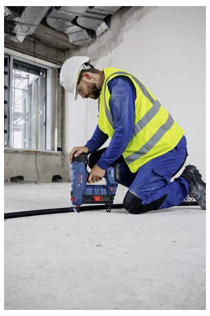 Robotnik budowlany w kamizelce odblaskowej i kasku roboczym klęczy na betonowej podłodze, używając elektronarzędzia w częściowo ukończonym pomieszczeniu.