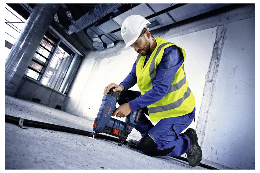 Robotnik budowlany w kamizelce odblaskowej i kasku używa elektronarzędzia na podłodze niedokończonego budynku.