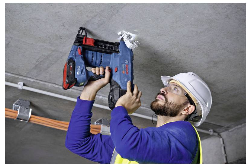 Robotnik budowlany w kasku ochronnym i odzieży roboczej używa elektronarzędzia do wiercenia w betonowym suficie, w pobliżu widoczne są przewody elektryczne.