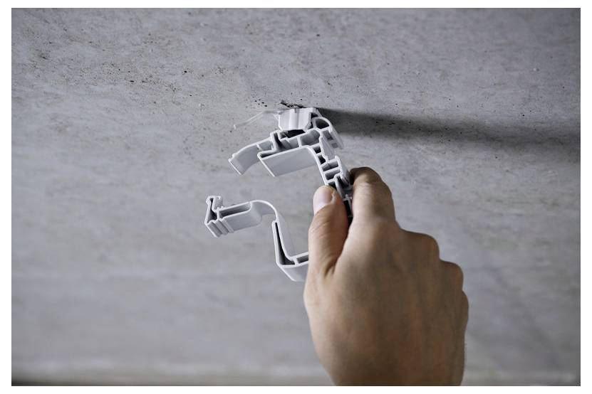 Ręka trzymająca szarą plastikową klamrę kablową na tle betonowej powierzchni, prawdopodobnie w celu instalacji lub naprawy.